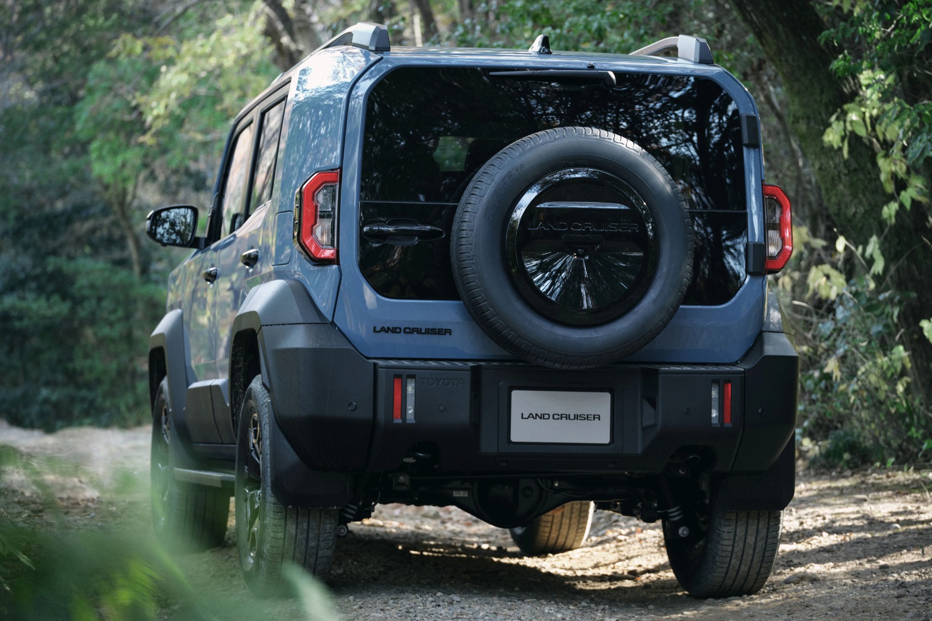 Blue Toyota Land Cruiser SUV with spare tire mounted on the rear door, parked on a dirt path in a forest.