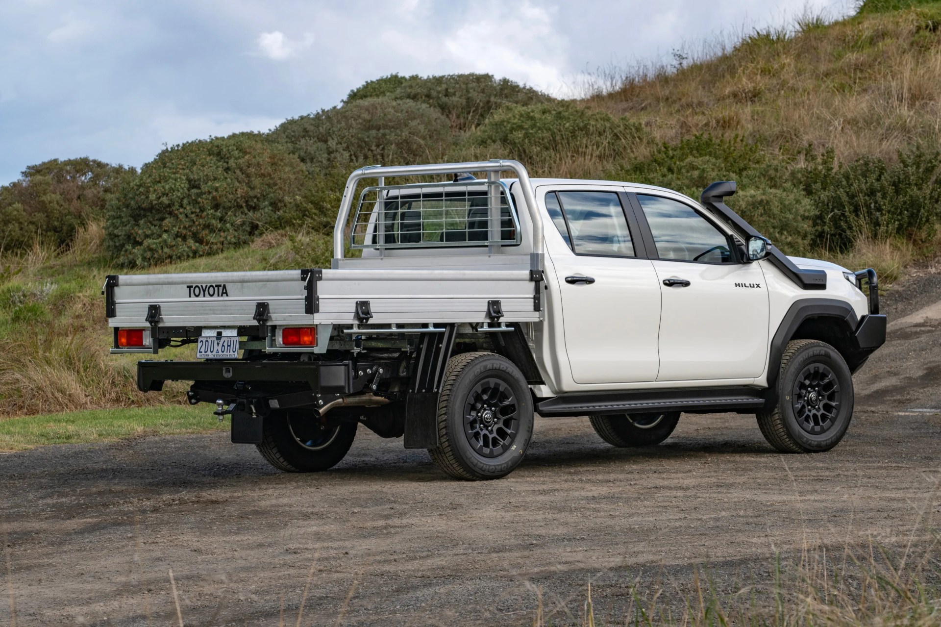 White Toyota Hilux pickup truck with black wheels and silver flatbed tray parked on a dirt road with grassy hills in the background.