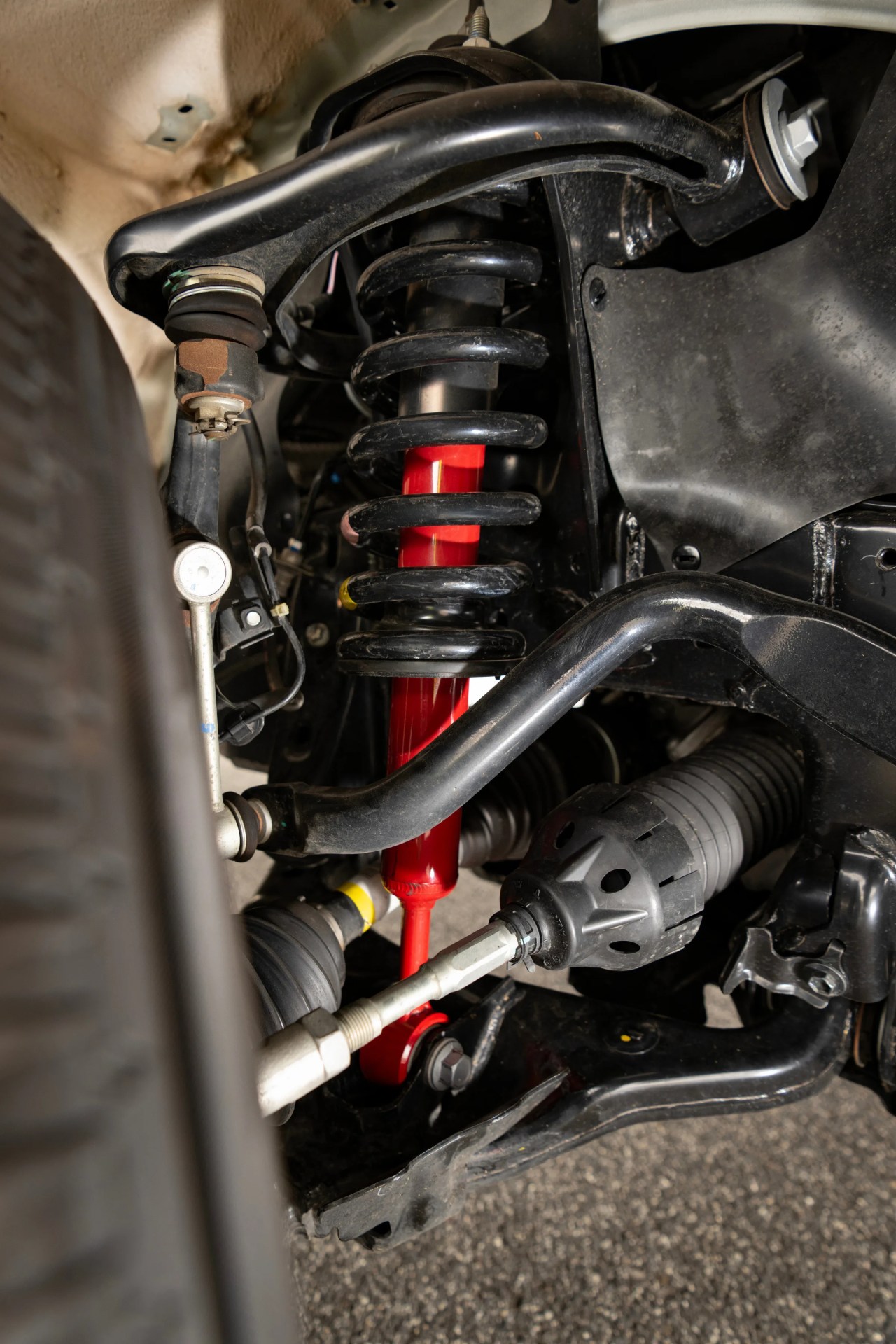 Close-up of a vehicle suspension system showing a black coil spring around a red shock absorber with surrounding black control arms and components.