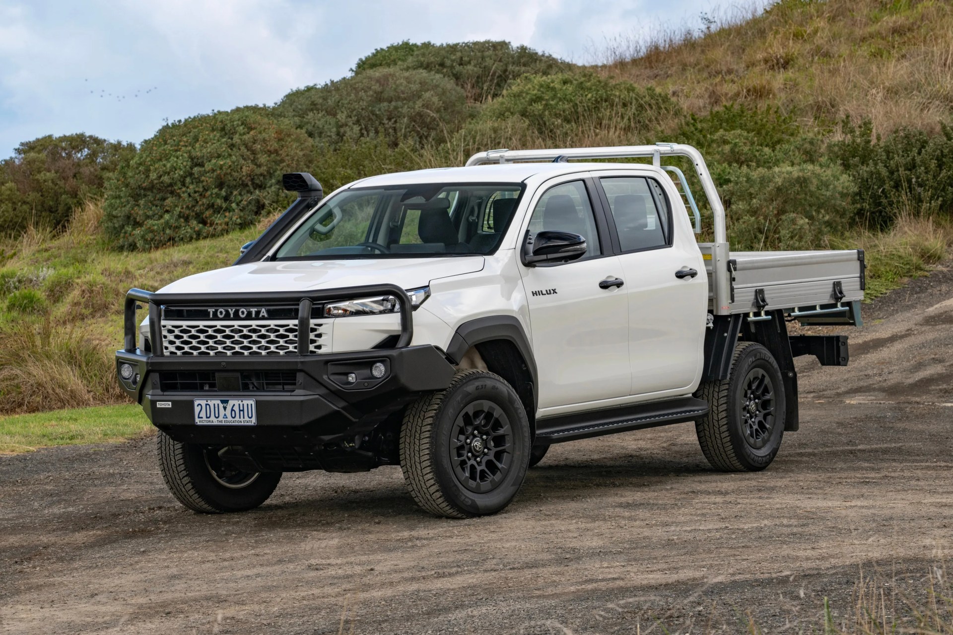 White Toyota Hilux pickup truck with black bull bar and snorkel parked on a dirt road with greenery in the background.