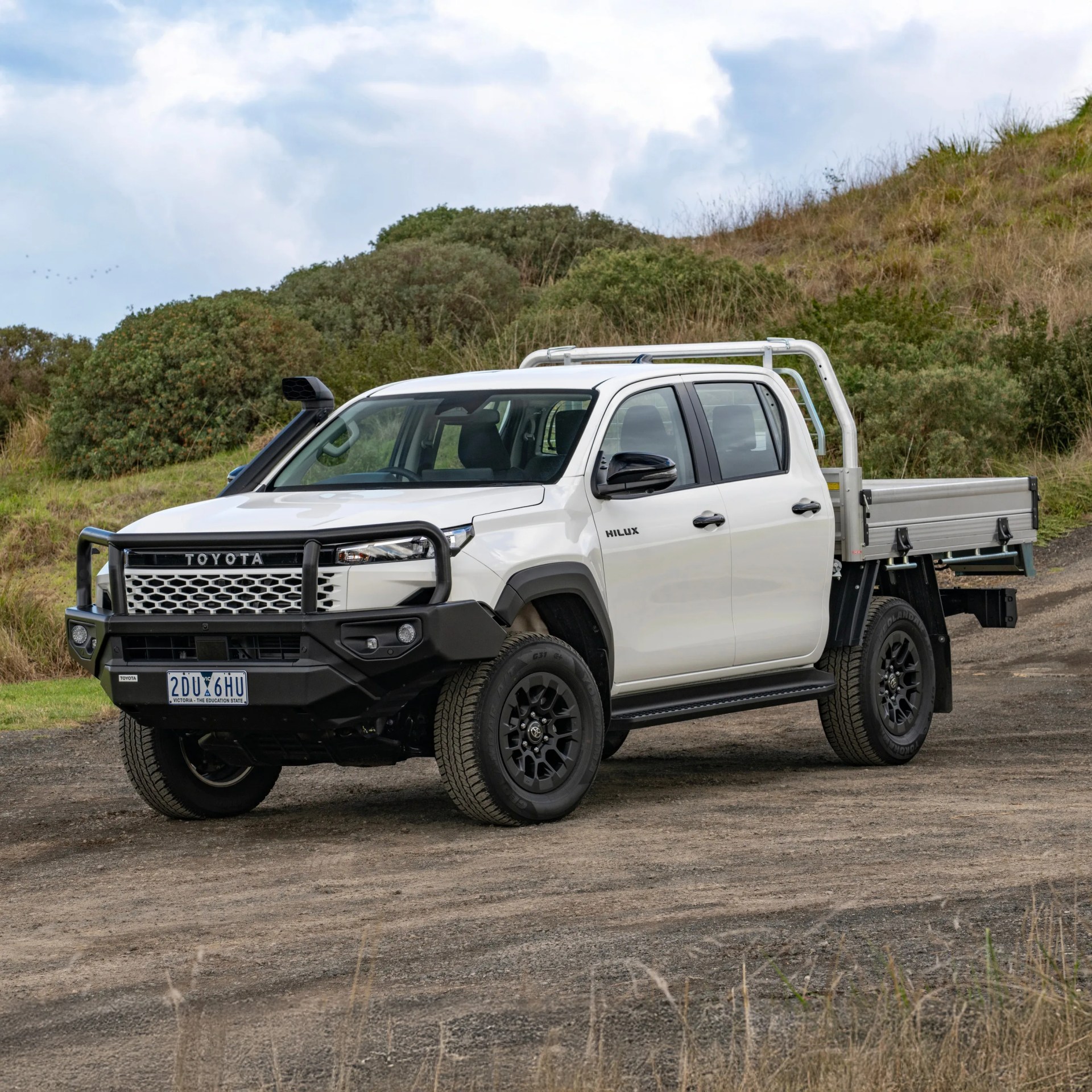 White Toyota Hilux pickup truck with black grille guard and snorkel parked on dirt road with green bushes in background.