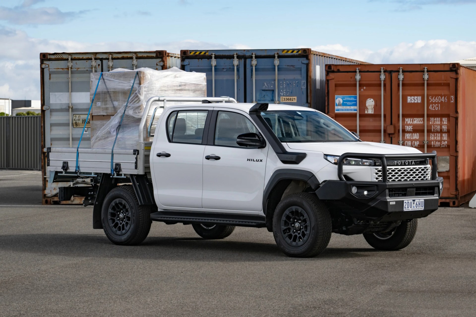 White Toyota Hilux pickup truck with black wheels and snorkel, carrying palletized cargo secured with blue straps, parked in front of shipping containers.