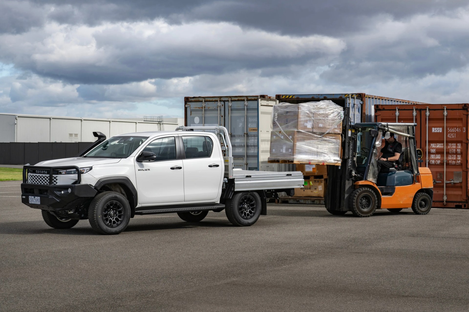 White Toyota Hilux pickup truck with black bull bar parked next to an orange forklift lifting wrapped pallets in an industrial area.