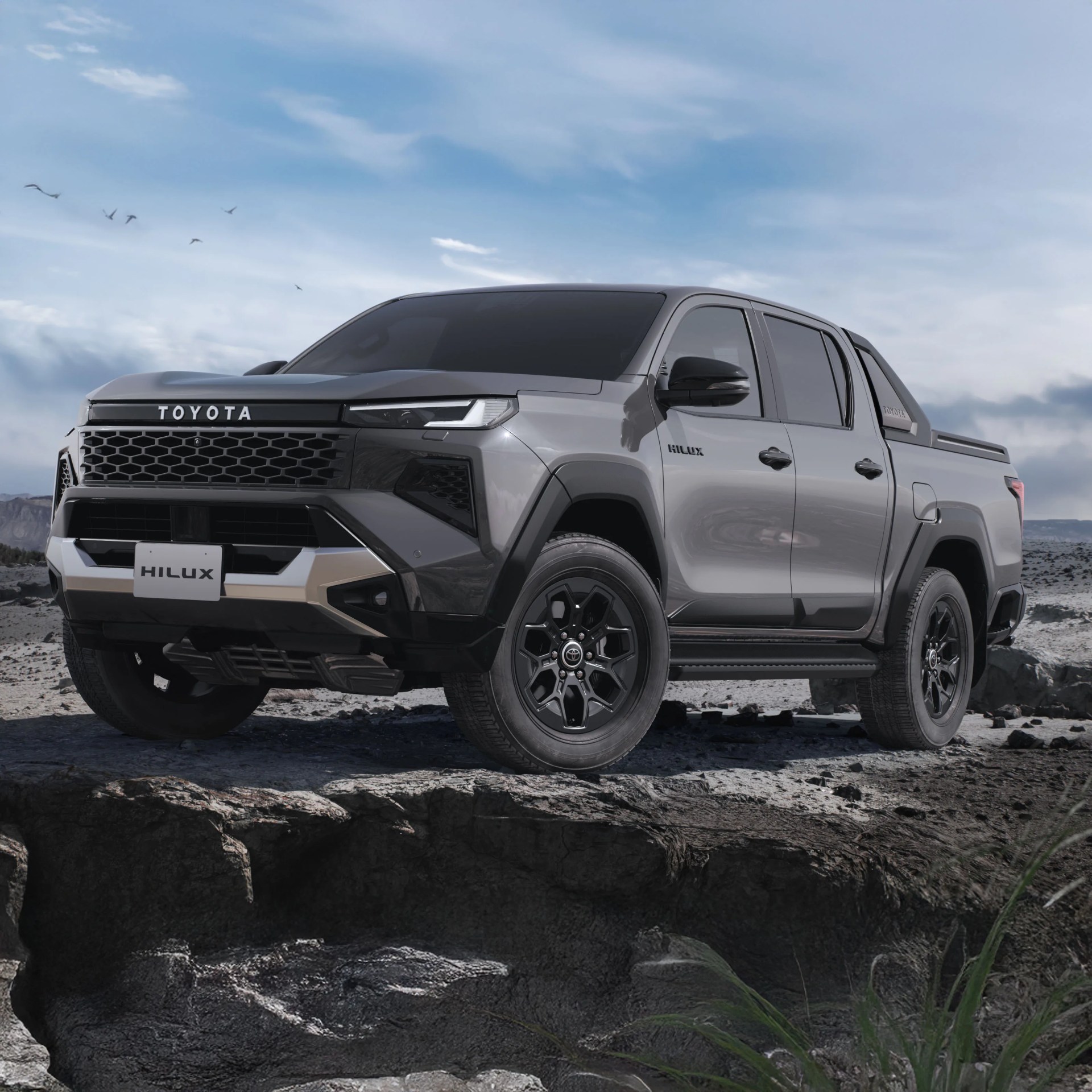 Gray Toyota Hilux pickup truck with black grille and wheels on rocky terrain under a cloudy sky.