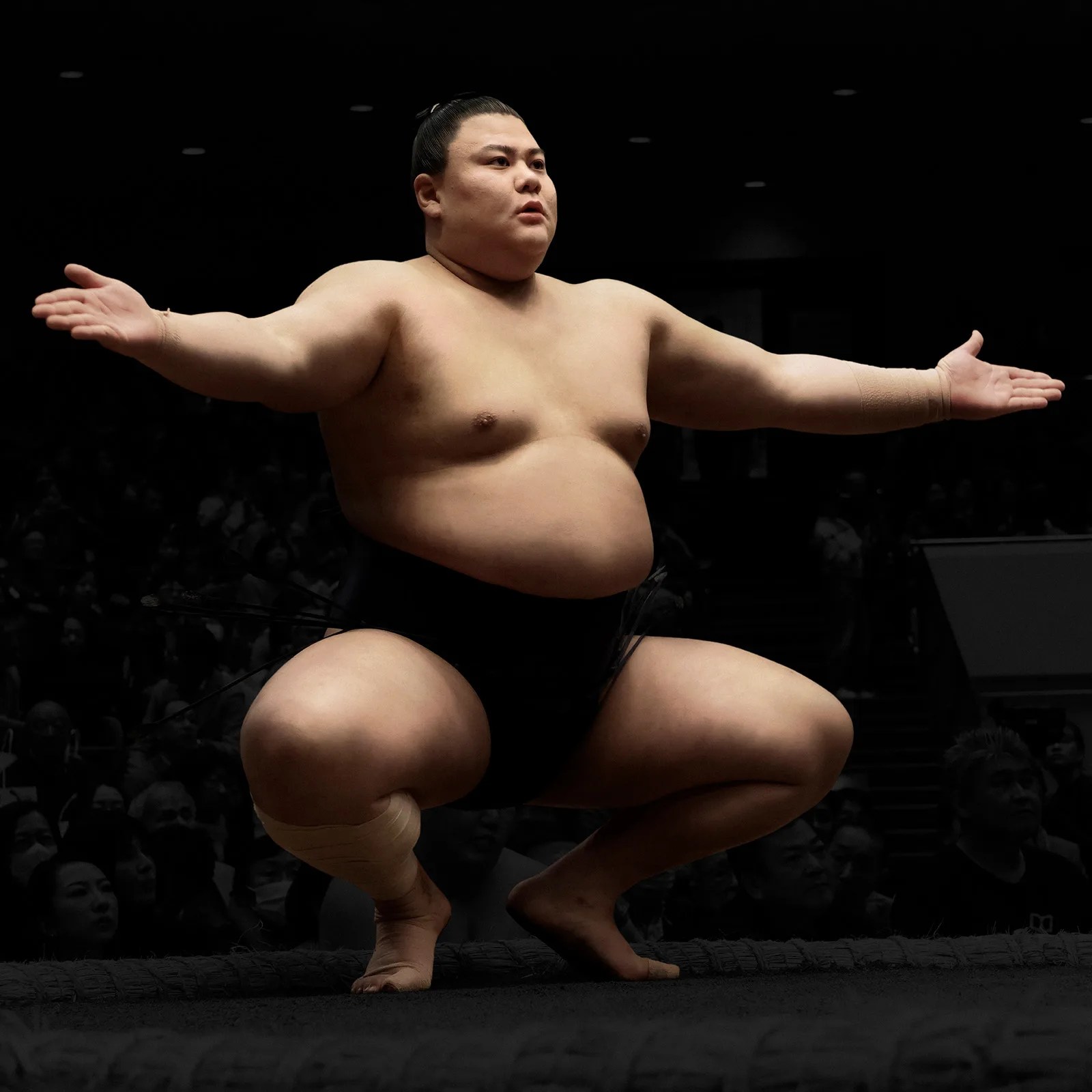 Sumo wrestler in black mawashi squatting with arms extended in a dark arena.