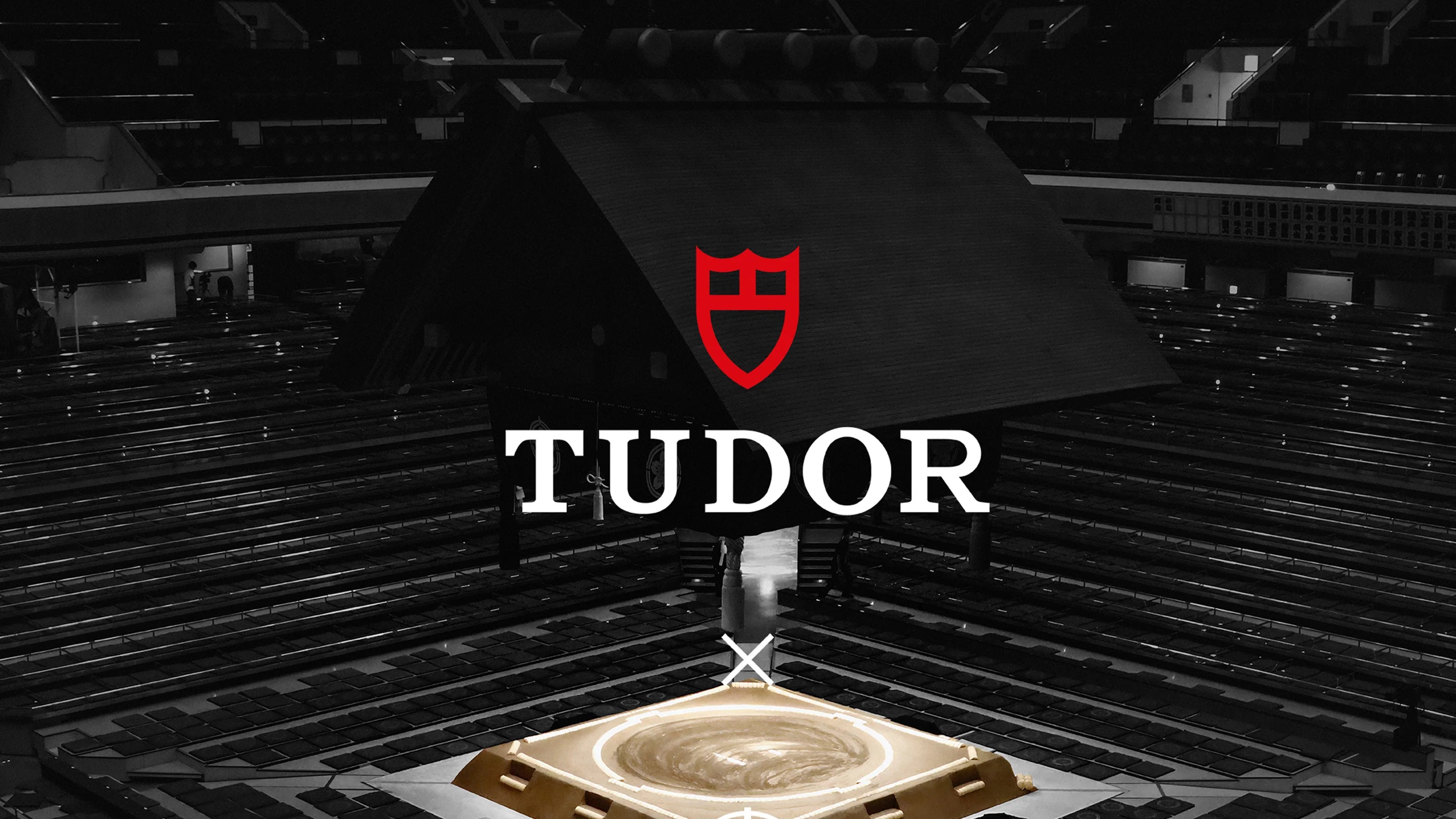 Sumo wrestling arena with a large wooden roof structure in the center and the Tudor logo overlayed.