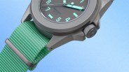 Close-up of a gray Unimatic watch with a green nylon strap and luminescent hands and numerals on a blue background.