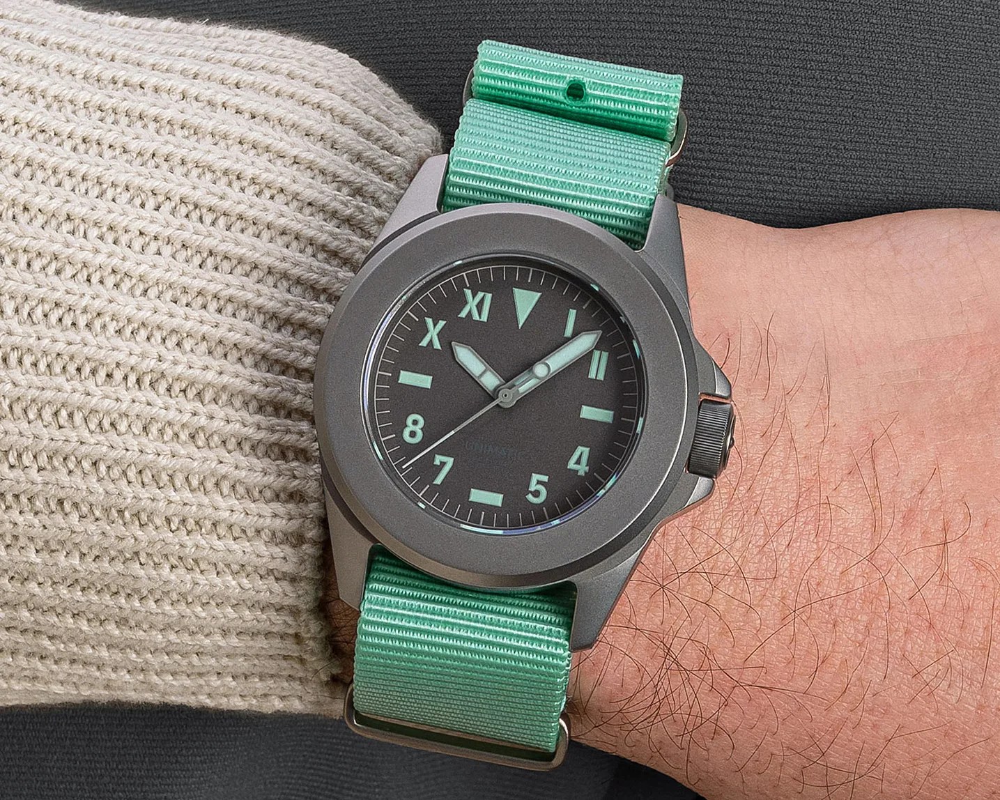 Wristwatch with a matte gray case, black dial with mint green luminous markers, and a mint green nylon strap worn on a wrist.