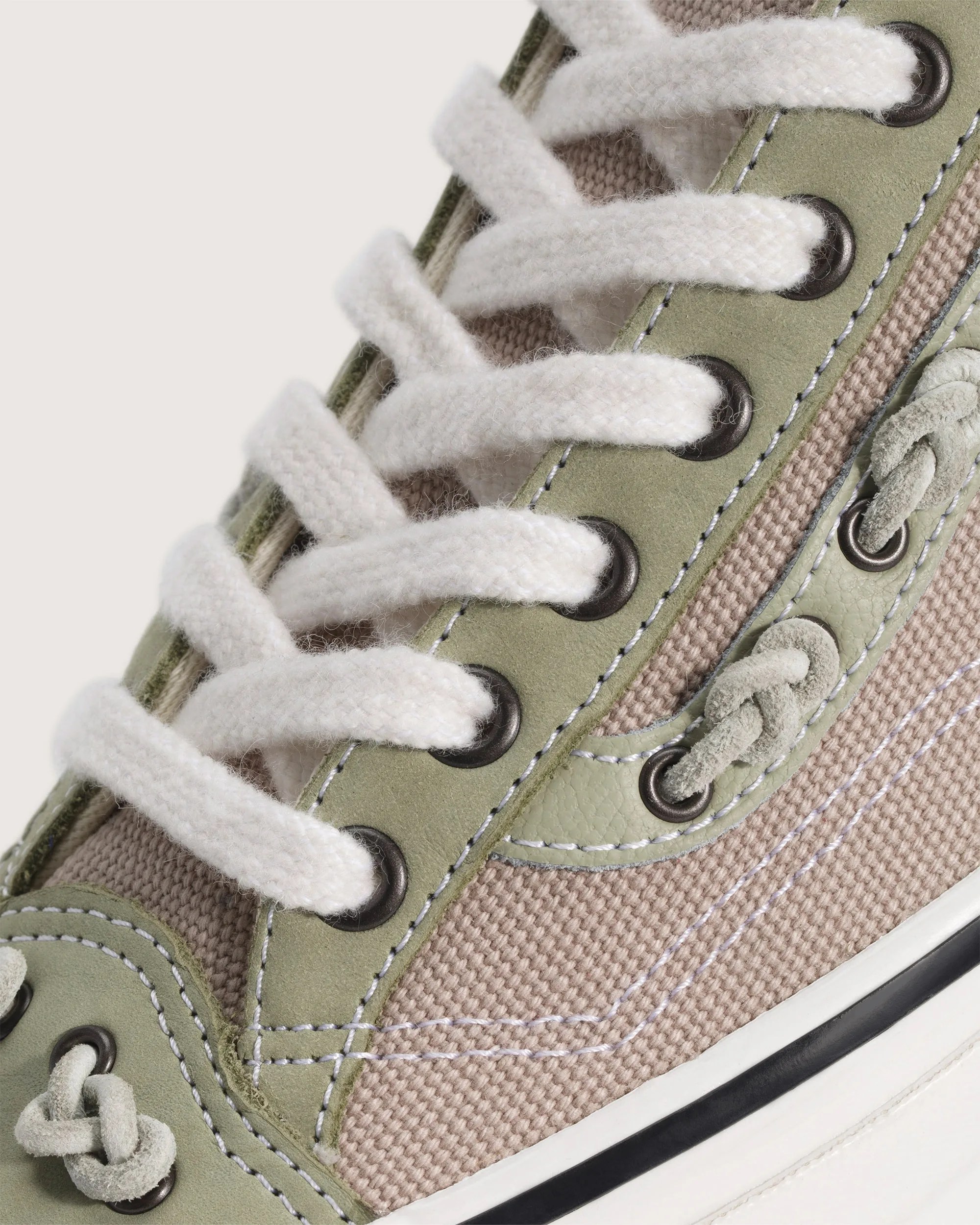 Close-up of beige canvas and olive green suede sneaker with white thick laces and black metal eyelets.