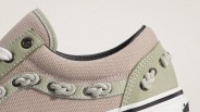 Close-up of beige canvas sneaker with olive green suede accents, white braided laces, and black eyelets.