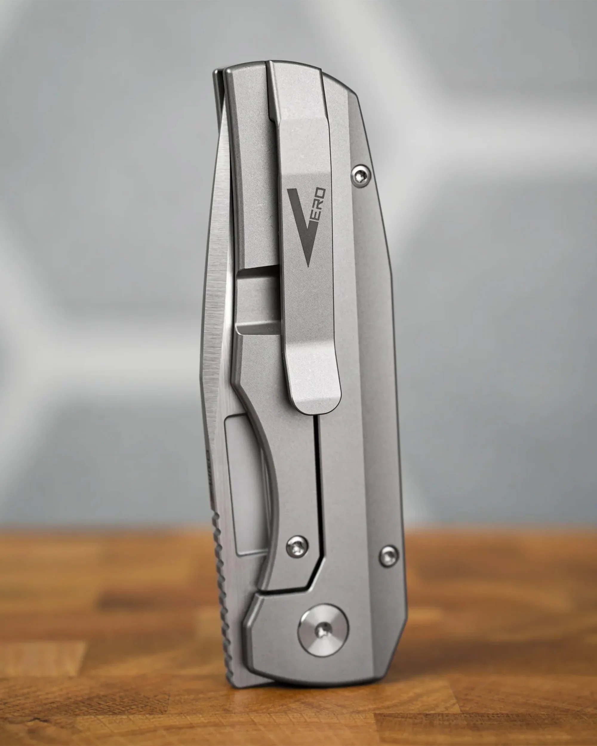 Closed silver folding knife with a clip labeled "Vero" standing on a wooden surface.