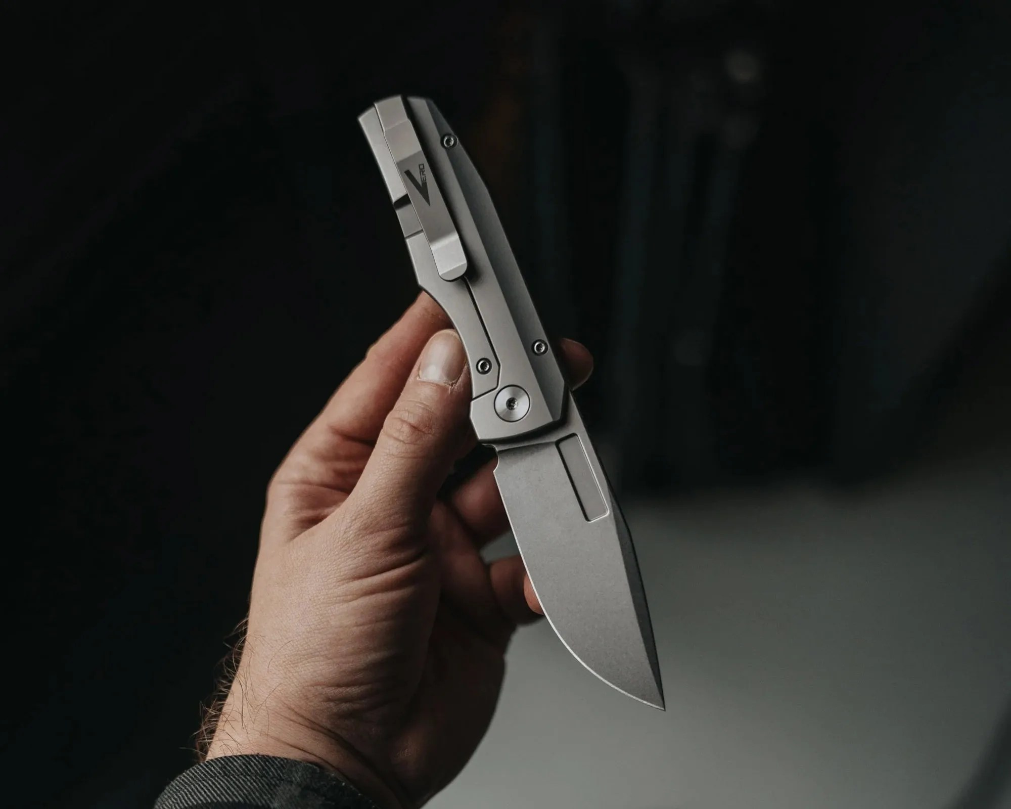 Hand holding a closed silver folding knife with a clip and visible screws on the handle.