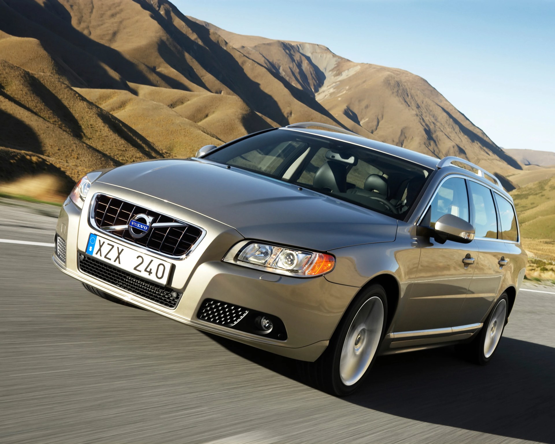 Silver Volvo station wagon driving on a road with dry brown hills in the background.