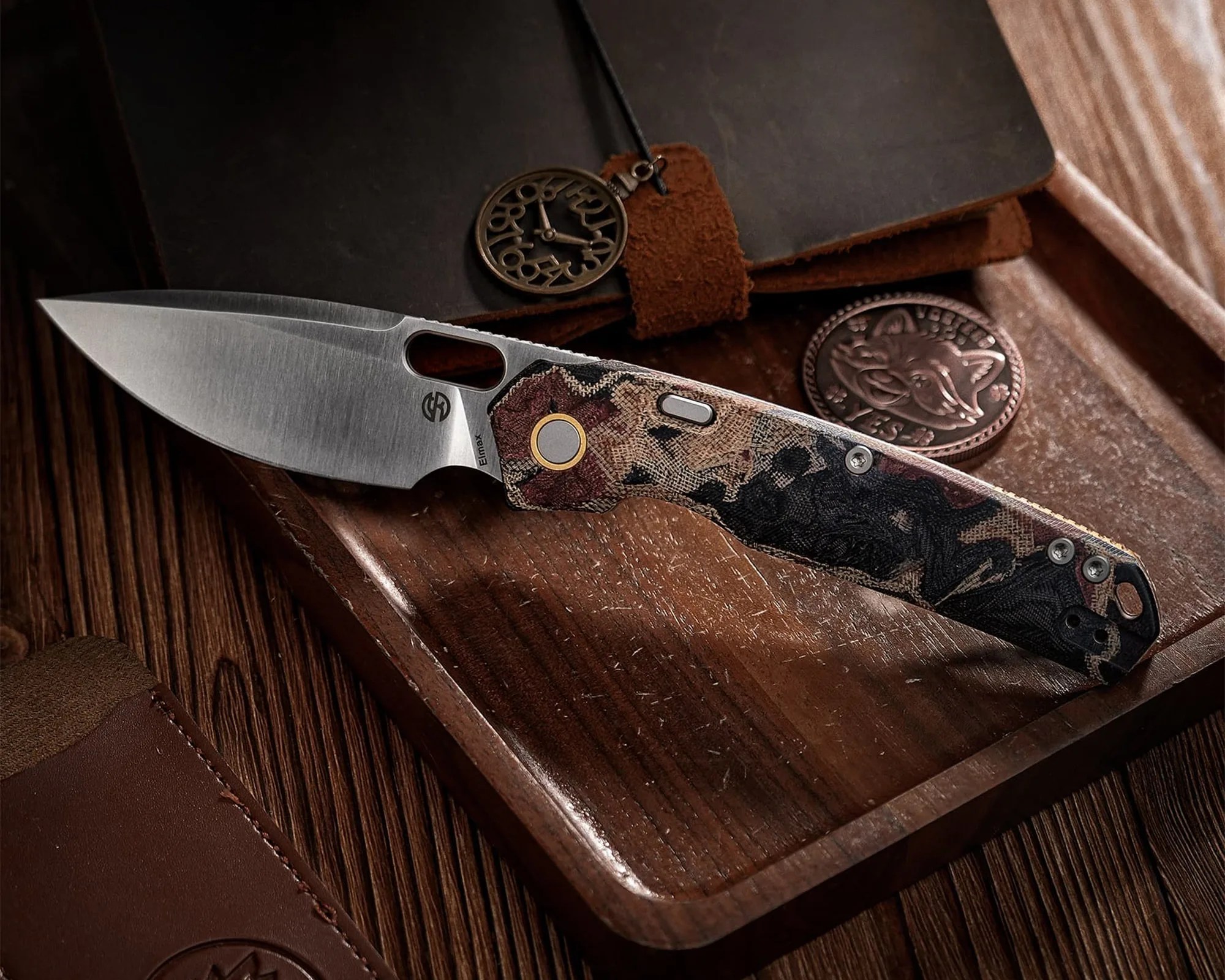 Folding knife with patterned handle and stainless steel blade resting on a wooden tray next to a leather notebook and round metal tokens.