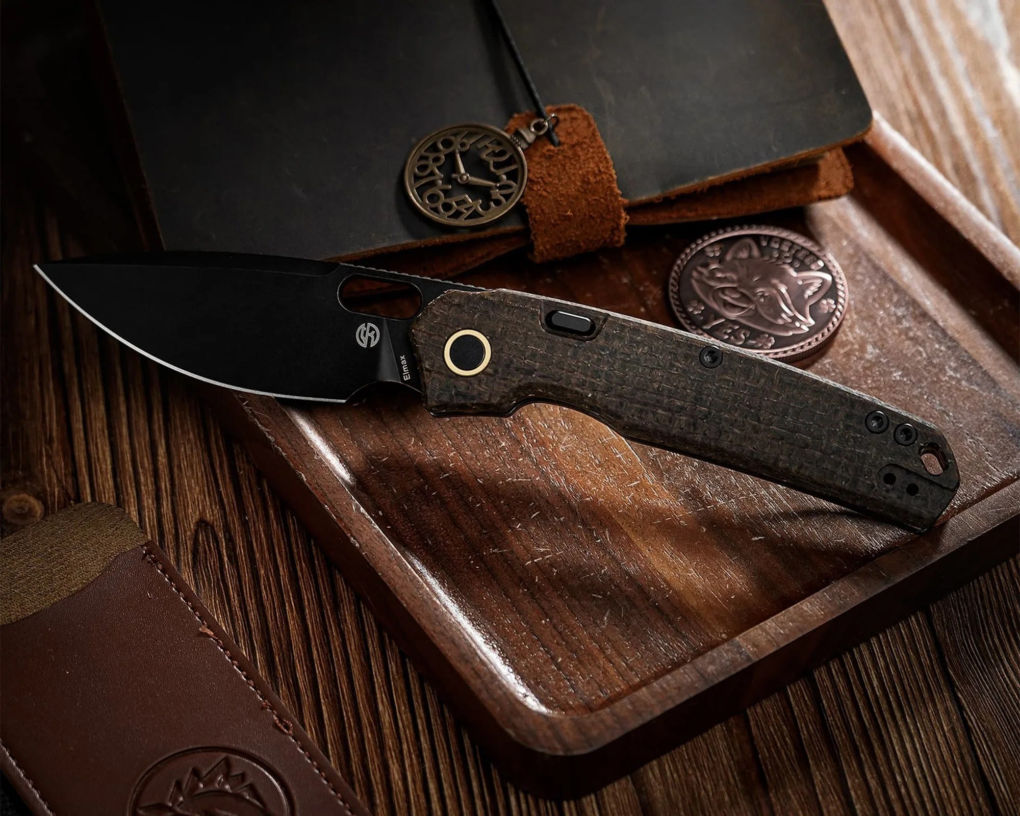 Black folding knife with textured handle on a wooden tray next to a leather notebook and a round coin with a fox design.