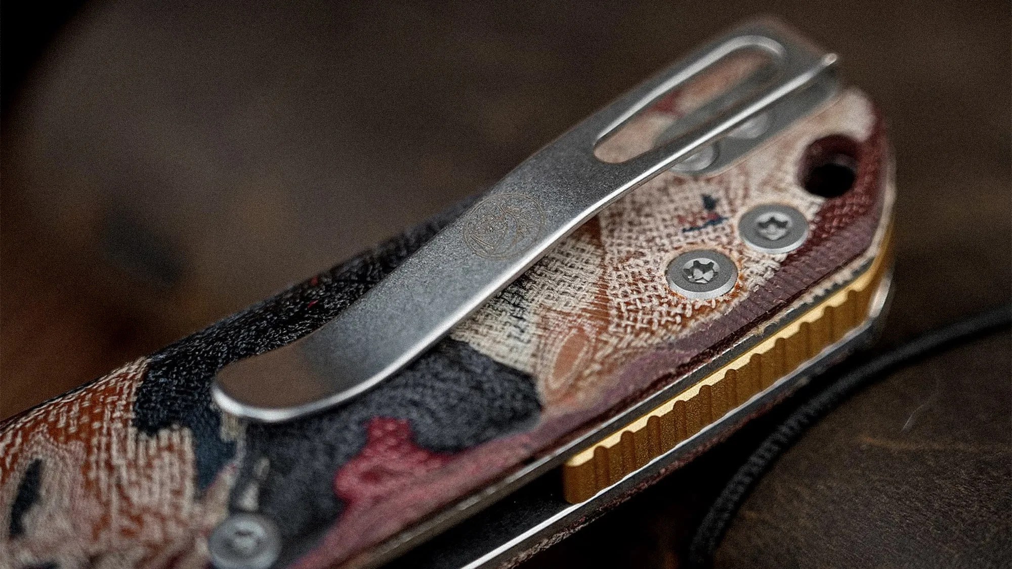 Close-up of a metal pocket clip attached to a textured, multicolored fabric handle with visible screws.