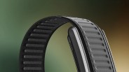 Close-up of a black textured watch strap with a silver smartwatch case against a blurred green and brown background.