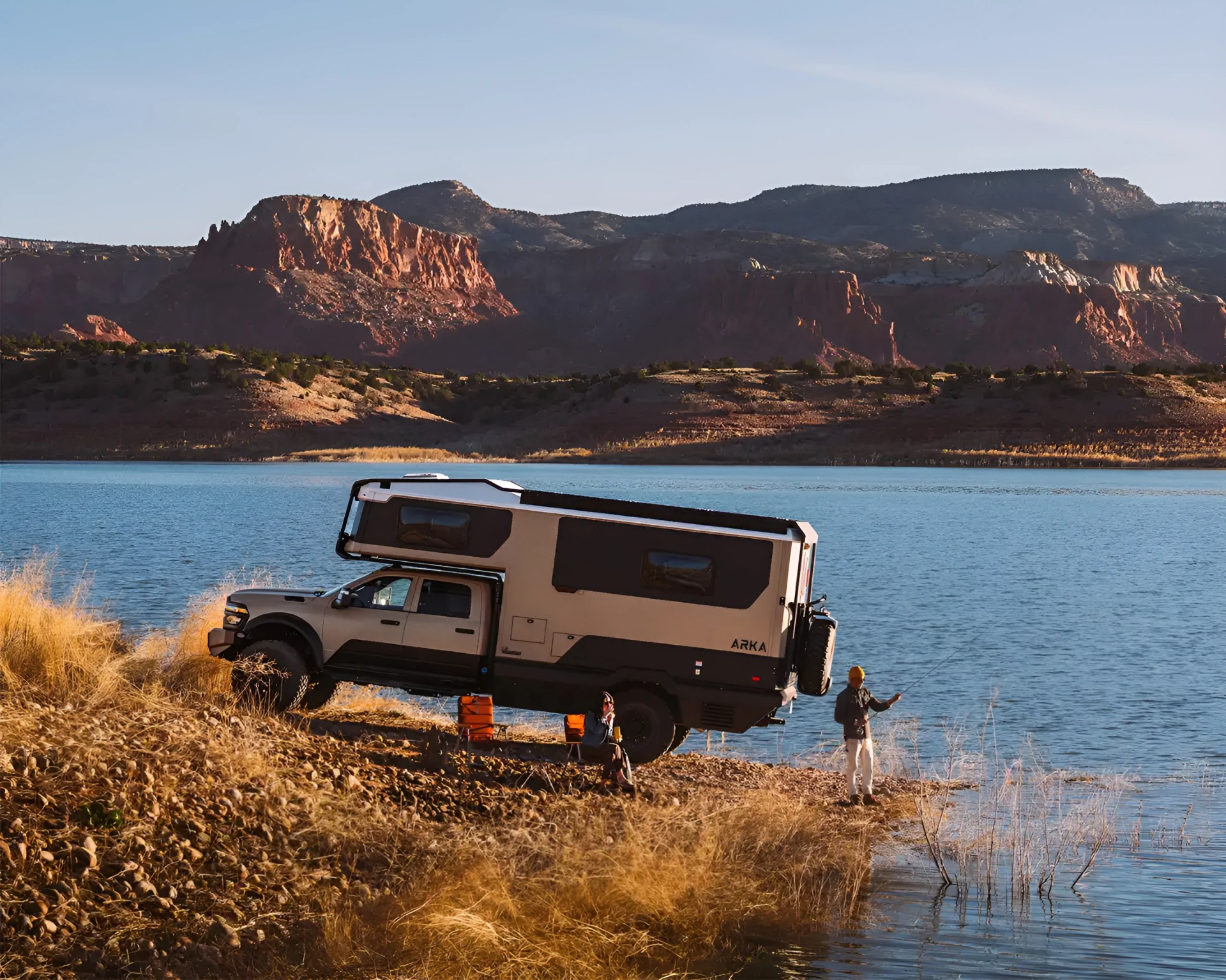 Beige and black ARKA camper truck parked by a lake with two people, one fishing and one sitting in a chair.