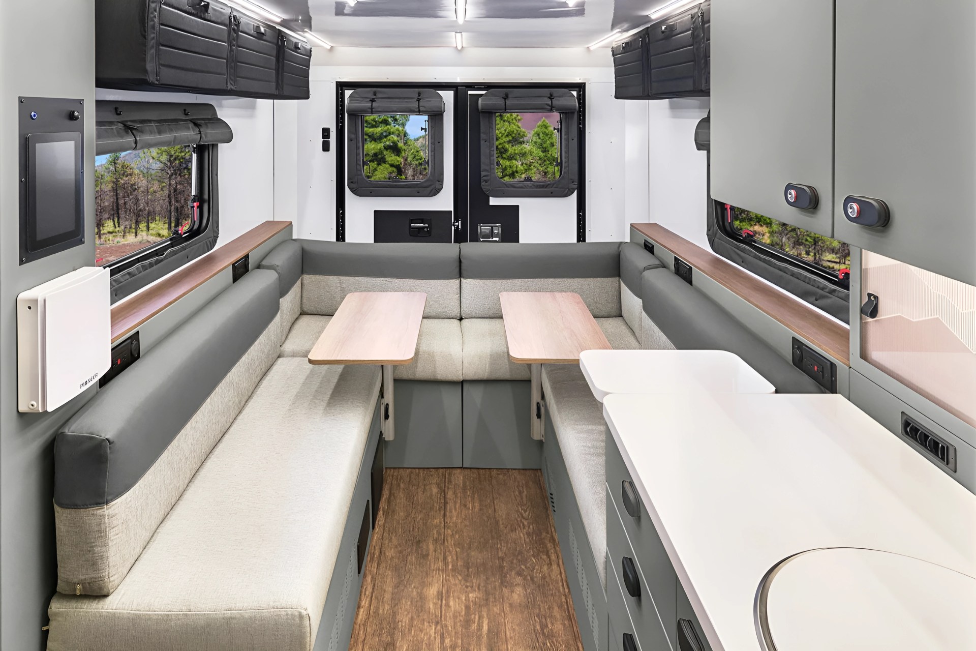 Compact camper interior with gray cushioned benches, two small wooden tables, overhead storage, and a white kitchenette counter.