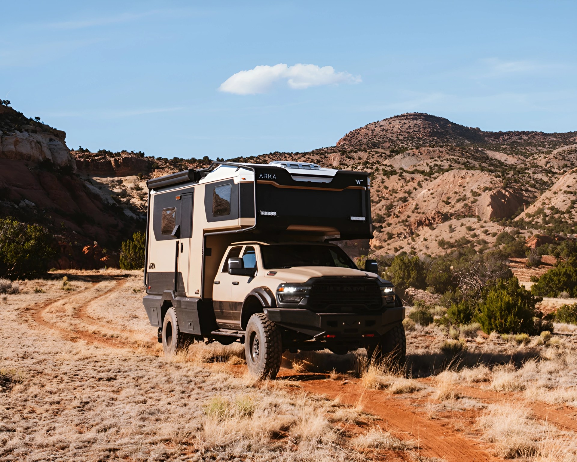 Beige and black off-road camper truck parked on a dirt trail in a rocky desert landscape.