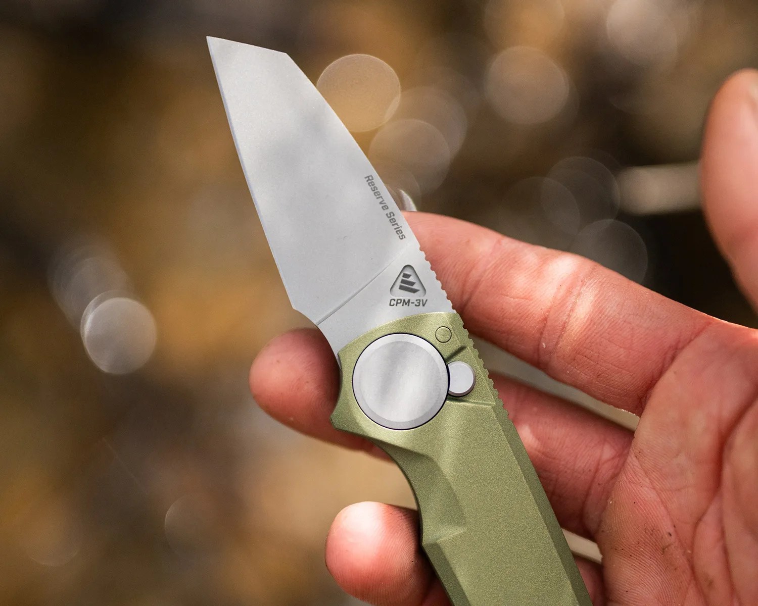 Hand holding a folding knife with a silver CPM-3V blade and olive green handle.