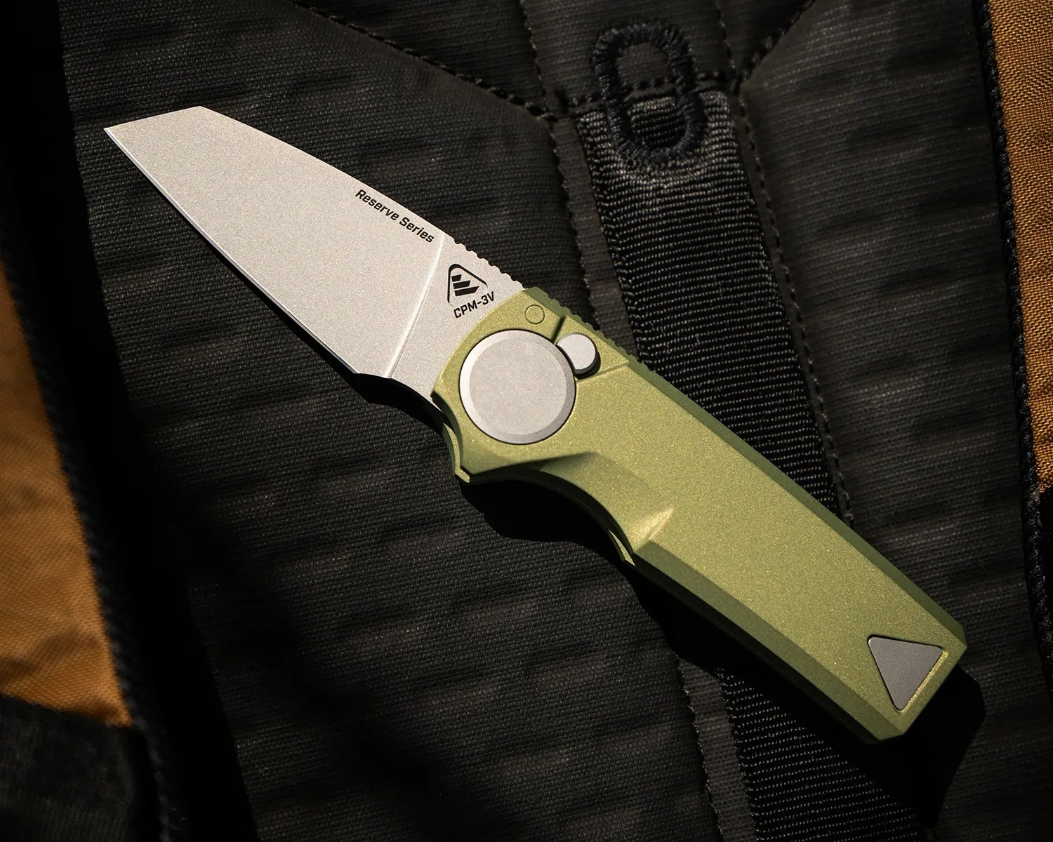 Folding knife with a stonewashed CPM-3V blade and olive green handle on black fabric.