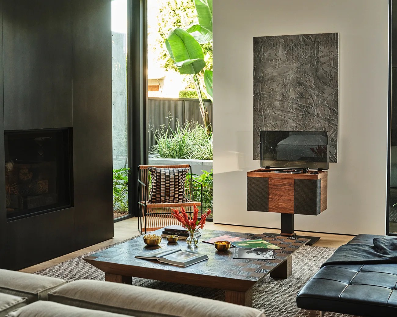 Wooden record player with black speaker panels and a transparent dust cover on a black pedestal in a modern living room.