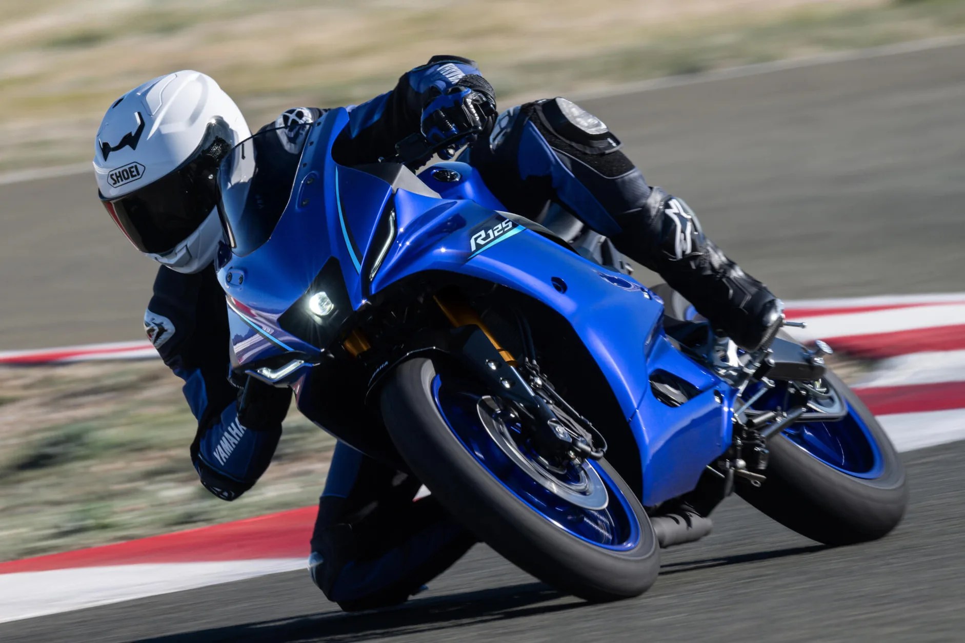 Blue Yamaha R125 sport motorcycle leaning sharply on a racetrack with rider in white Shoei helmet and black gear.