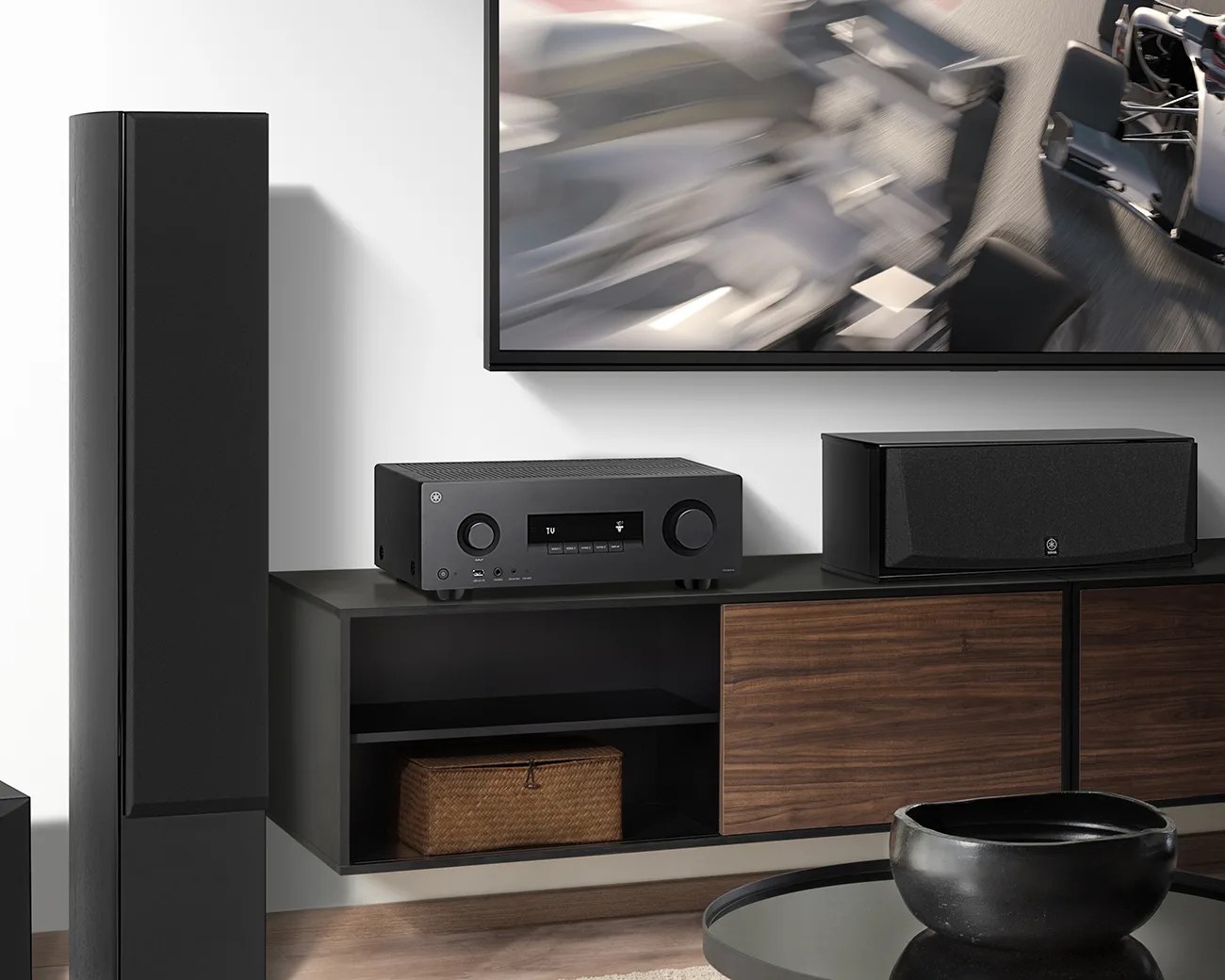 Black home audio receiver and center speaker on a black and wood media console under a wall-mounted TV.