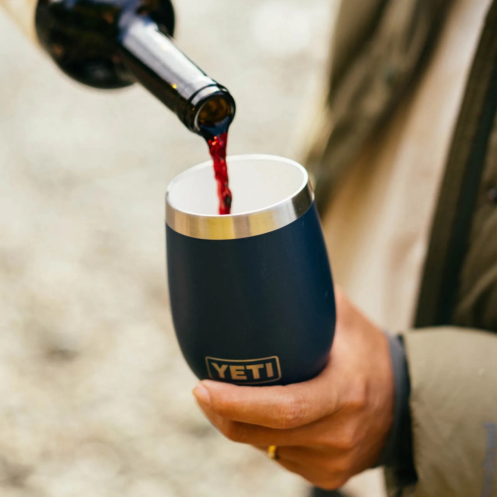 Red wine being poured from a dark bottle into a navy blue YETI tumbler held in a hand.