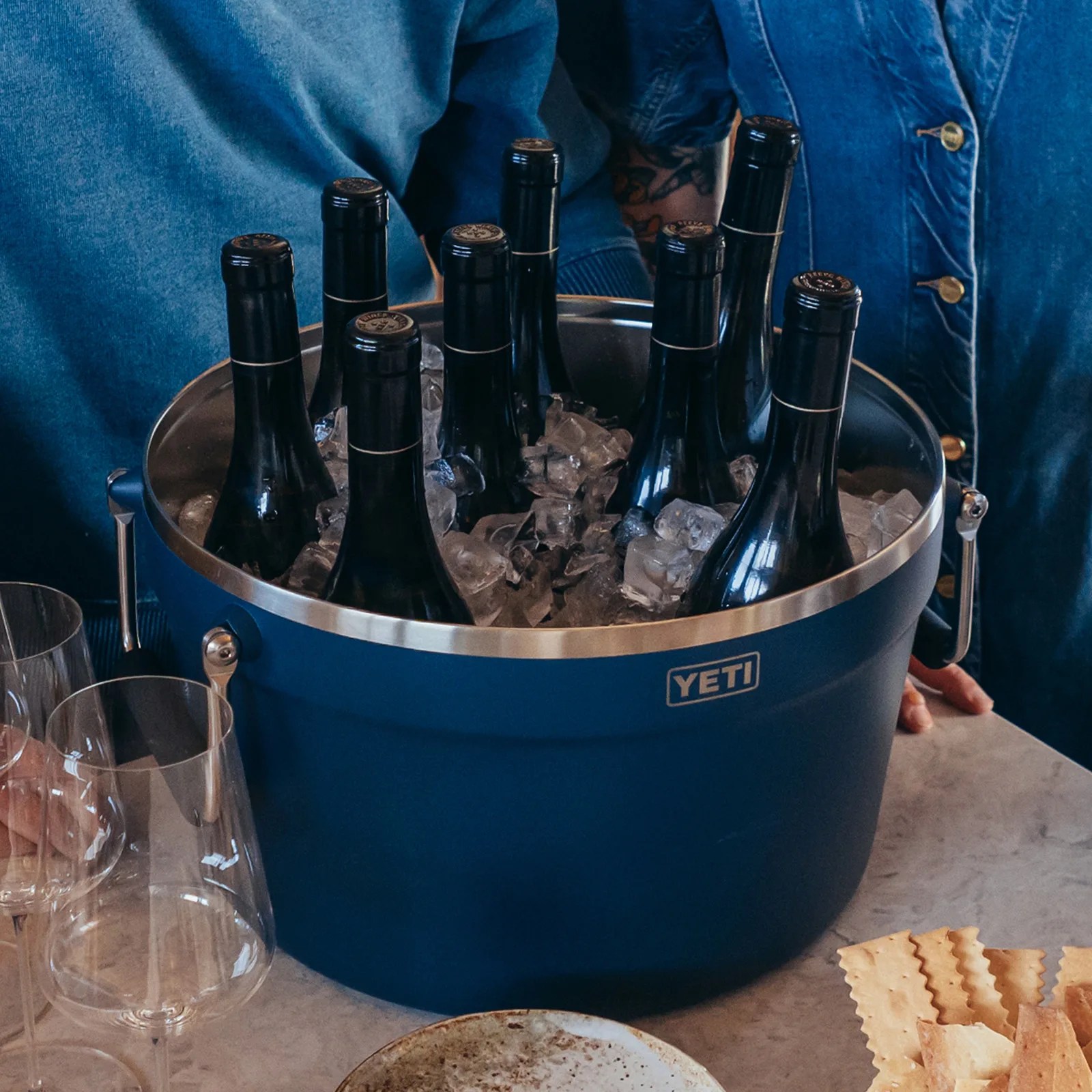 Blue YETI cooler filled with ice and nine black wine bottles on a table with glasses and crackers nearby.