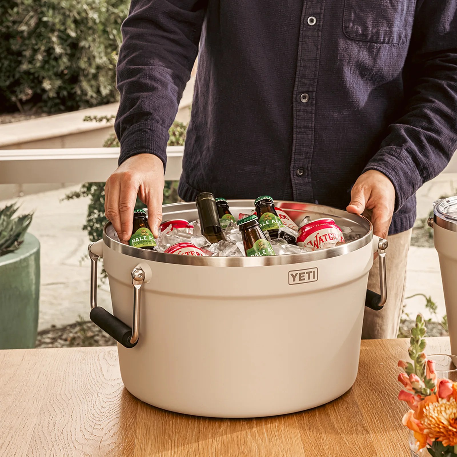 Beige YETI cooler bucket filled with ice, beer bottles, and soda cans held by a person in a dark shirt.
