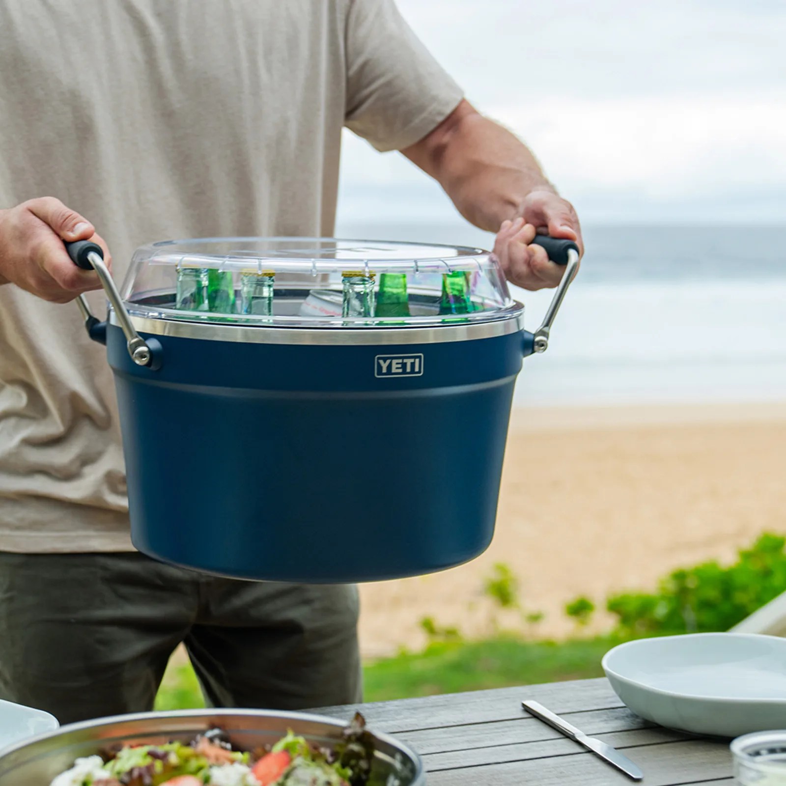 Blue YETI cooler with clear lid holding bottles and cans, carried by a person near a beach table.