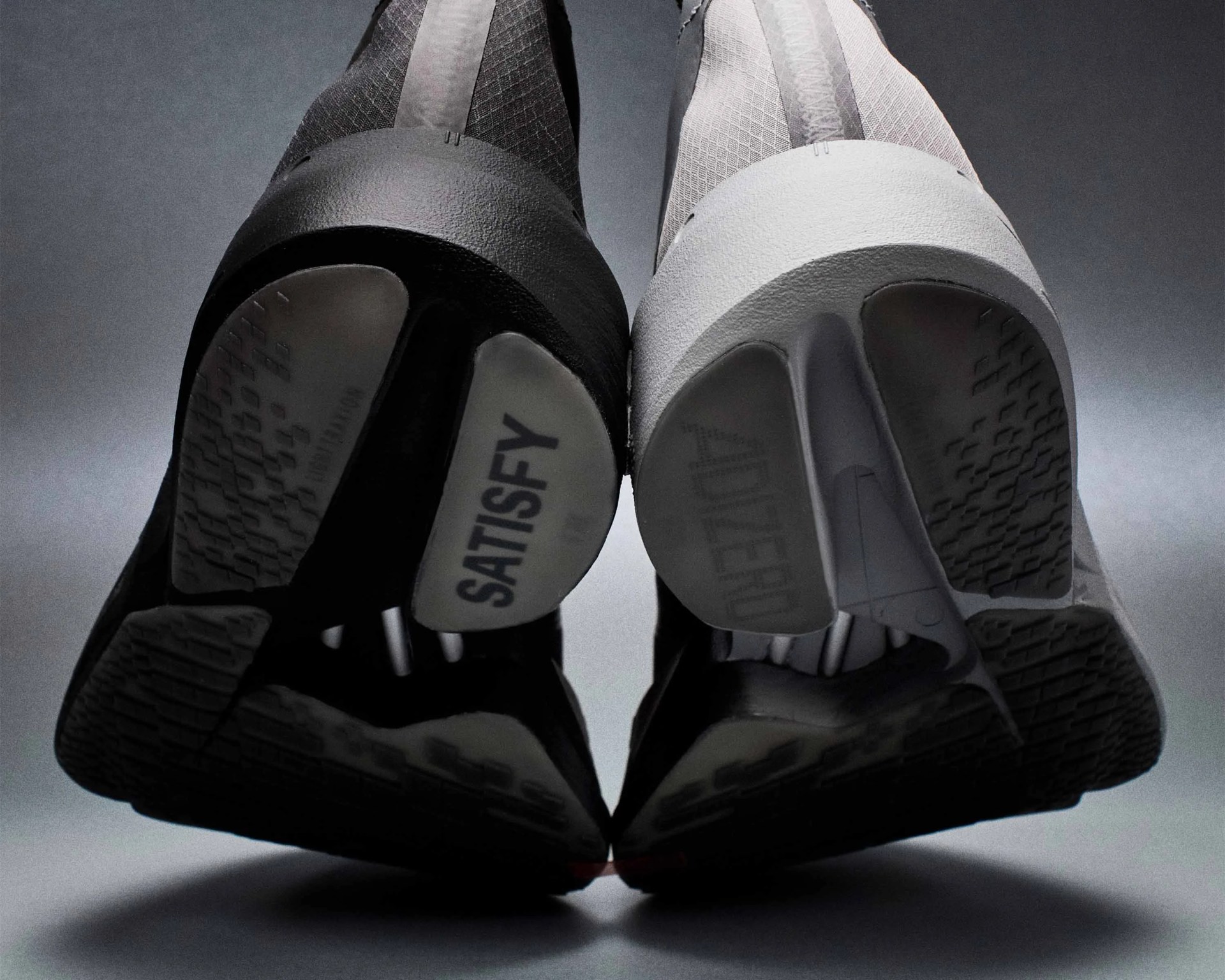 Pair of black and white sneakers with textured soles, one sole labeled "SATISFY," viewed from below.