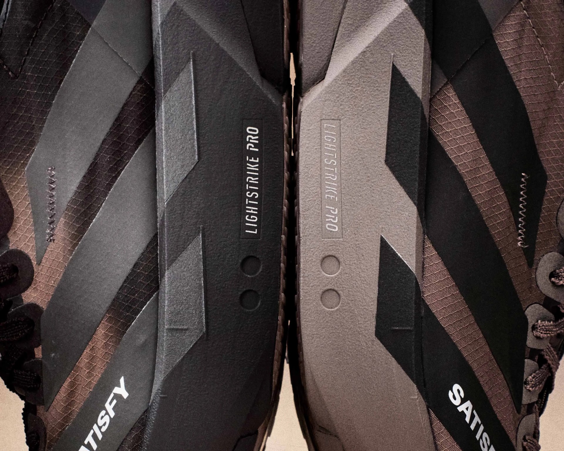 Close-up of two athletic shoes side by side, one black and one gray, both with "LIGHTSTRIKE PRO" text on the soles and "SATISFY" branding on the sides.