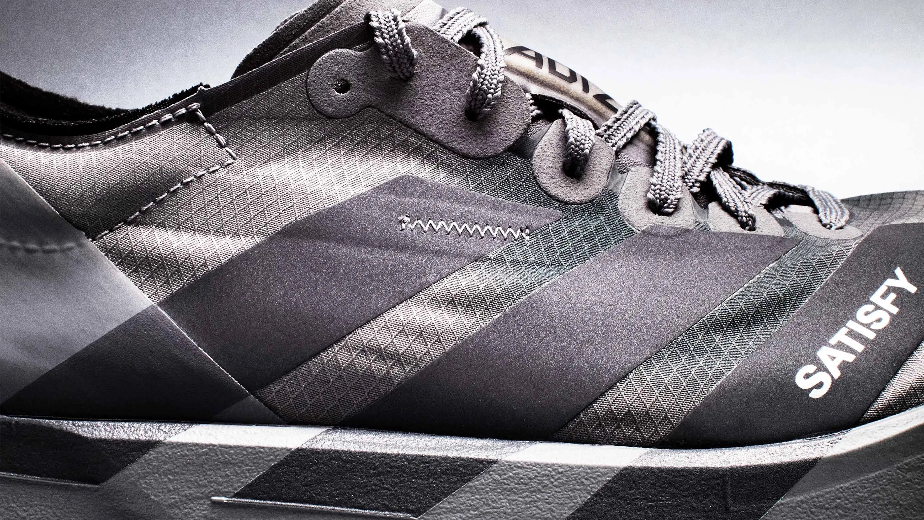 Close-up of a gray Adidas sneaker with textured fabric, braided laces, and the word "SATISFY" printed on the side.