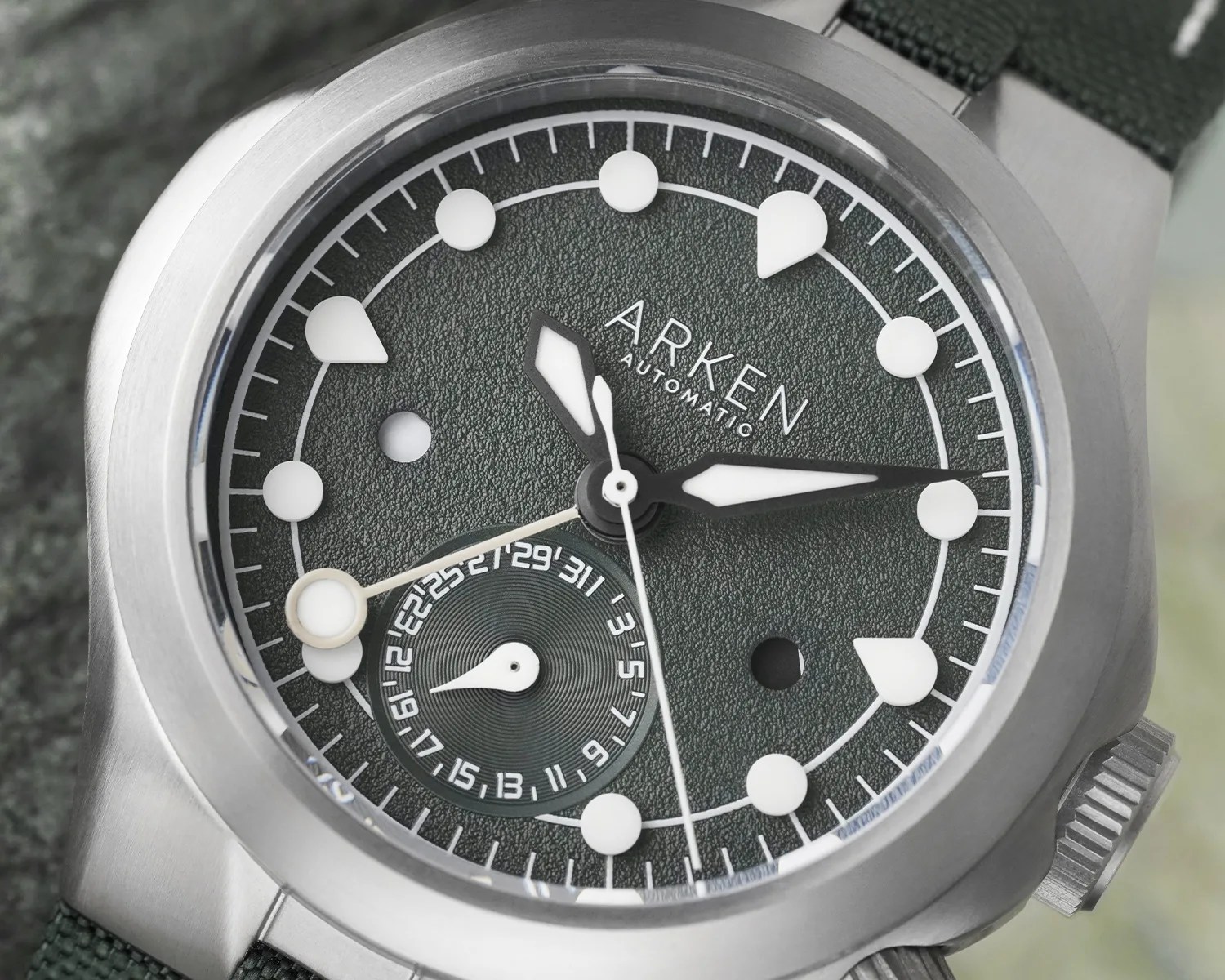 Close-up of an Arken Automatic watch with a textured dark green dial, white hour markers, and a date subdial.