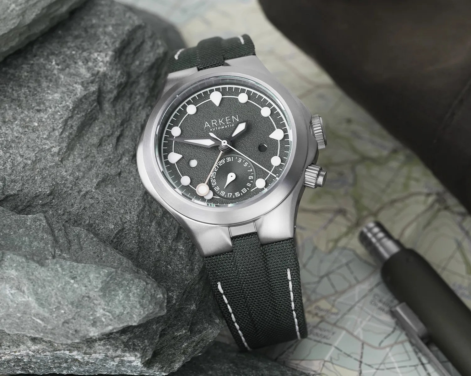 Titanium Arken automatic watch with textured dark gray dial and black fabric strap on rocks and map.