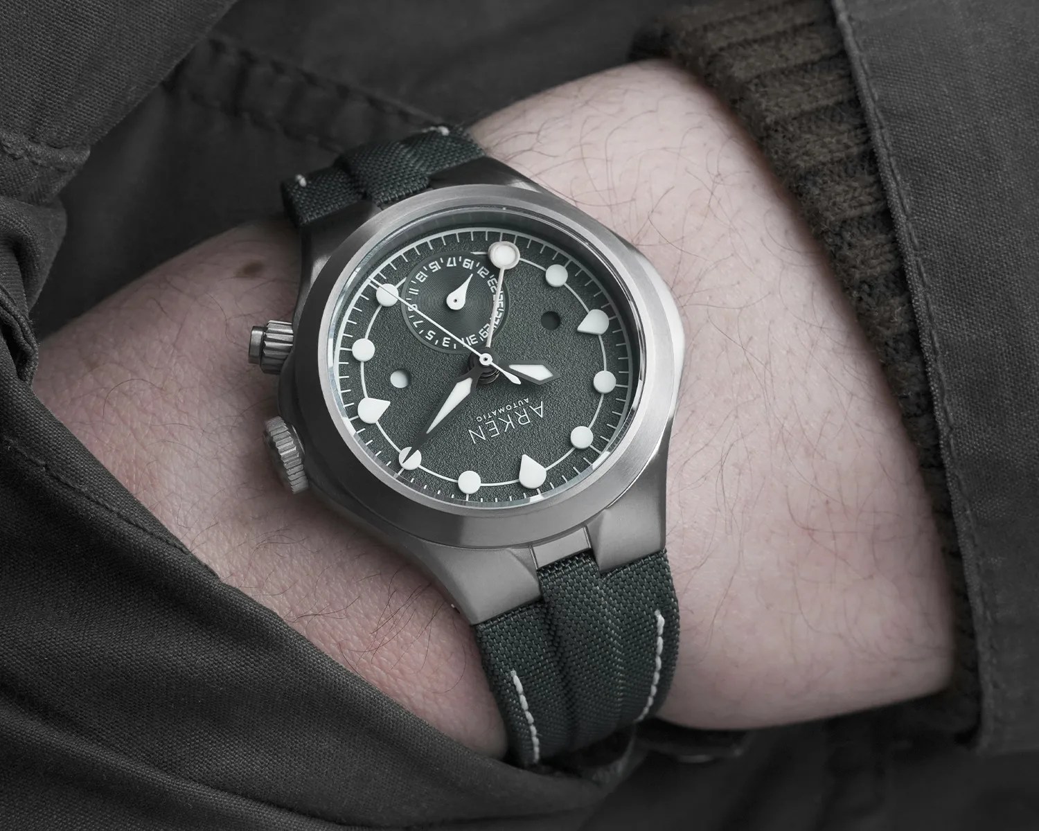 Matte silver Arken automatic wristwatch with textured dark green dial and matching fabric strap on a wrist.