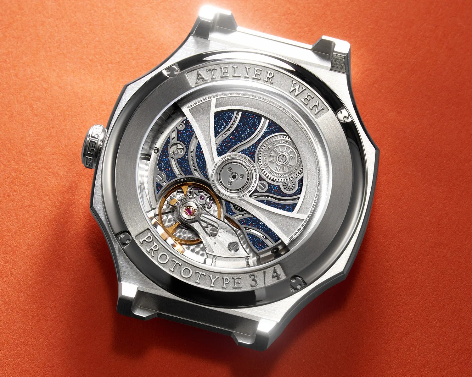 Silver Atelier Wen watch case back with visible intricate mechanical movement and blue speckled background on an orange surface.