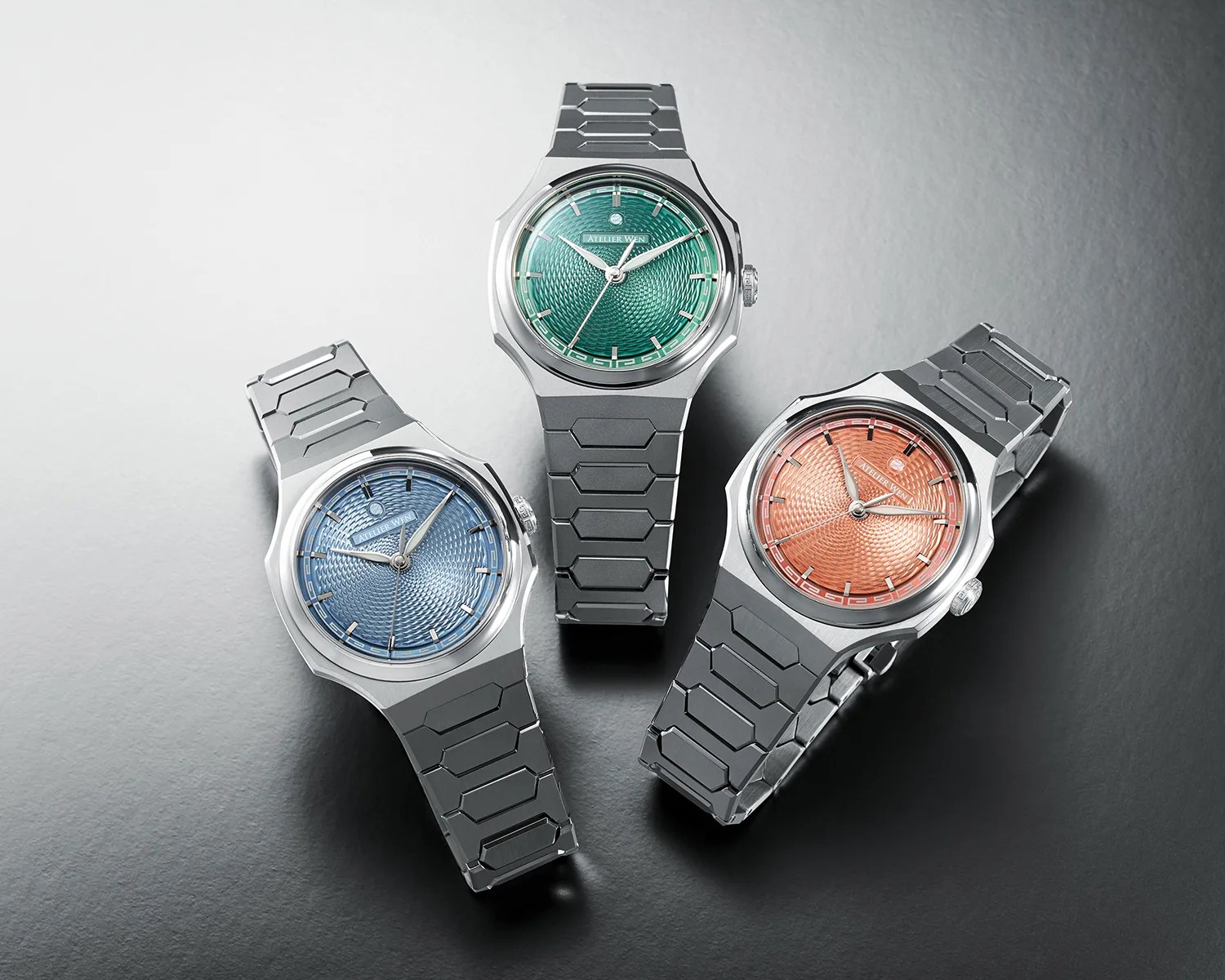 Three stainless steel wristwatches with textured dials in green, blue, and orange on a gray surface.