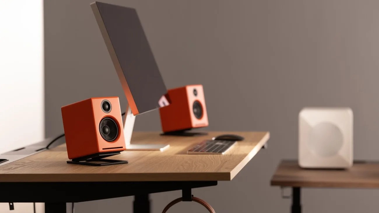 Pair of orange desktop speakers on black stands flanking a computer monitor on a wooden desk.