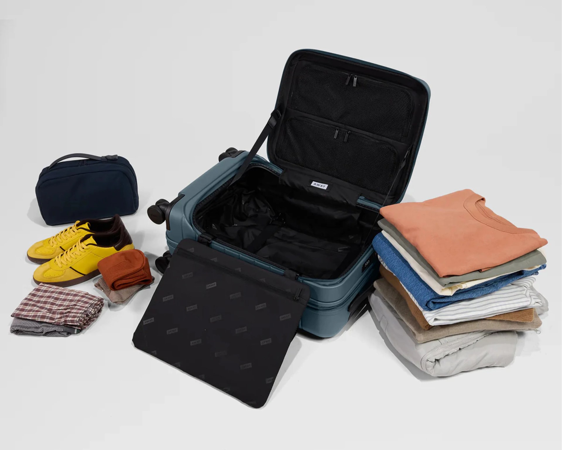 Open teal hard-shell suitcase with black interior and packing cubes, surrounded by folded clothes, yellow sneakers, and a navy toiletry bag.
