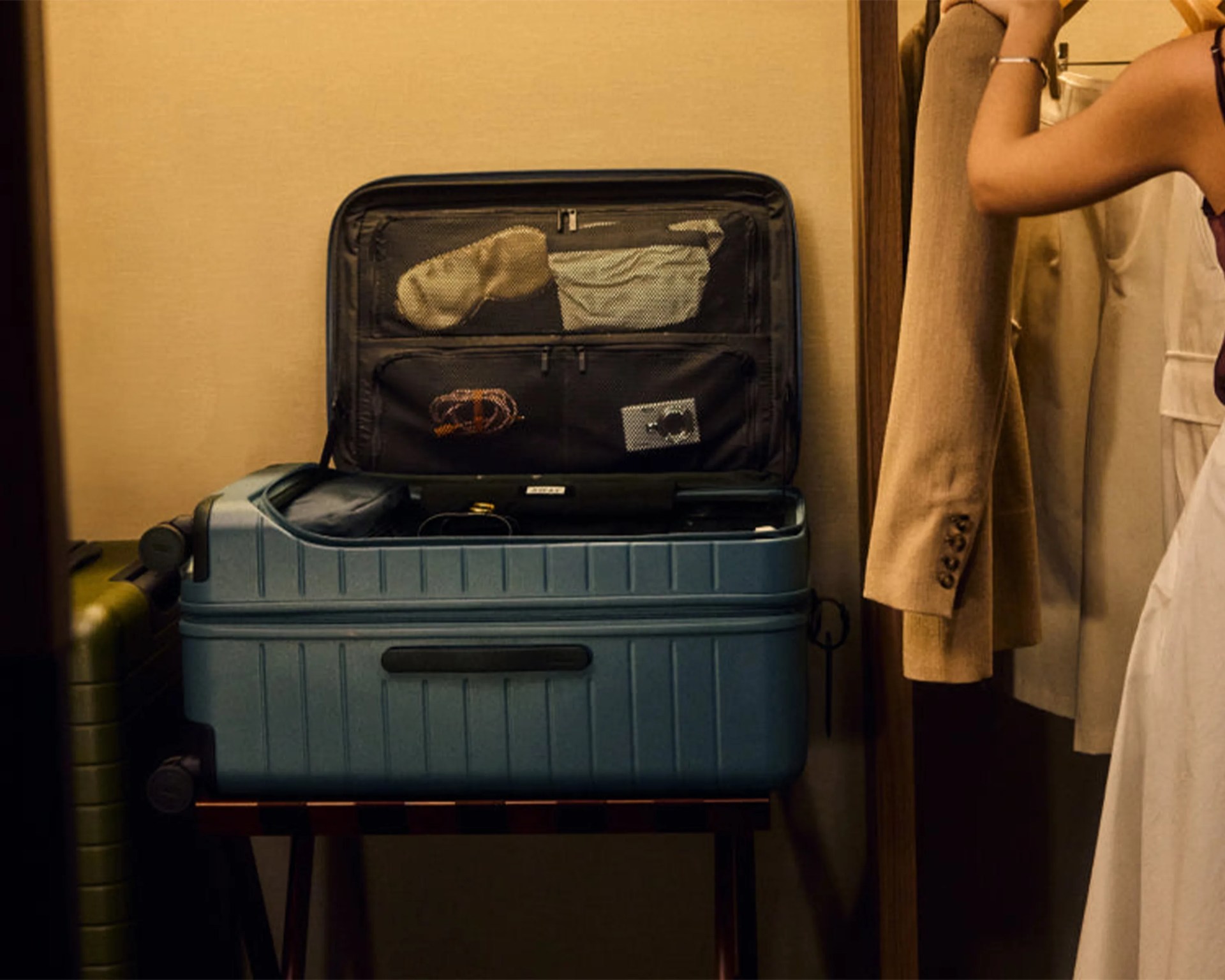 Blue hard-shell suitcase open with clothes inside on a luggage rack next to a beige coat hanging.