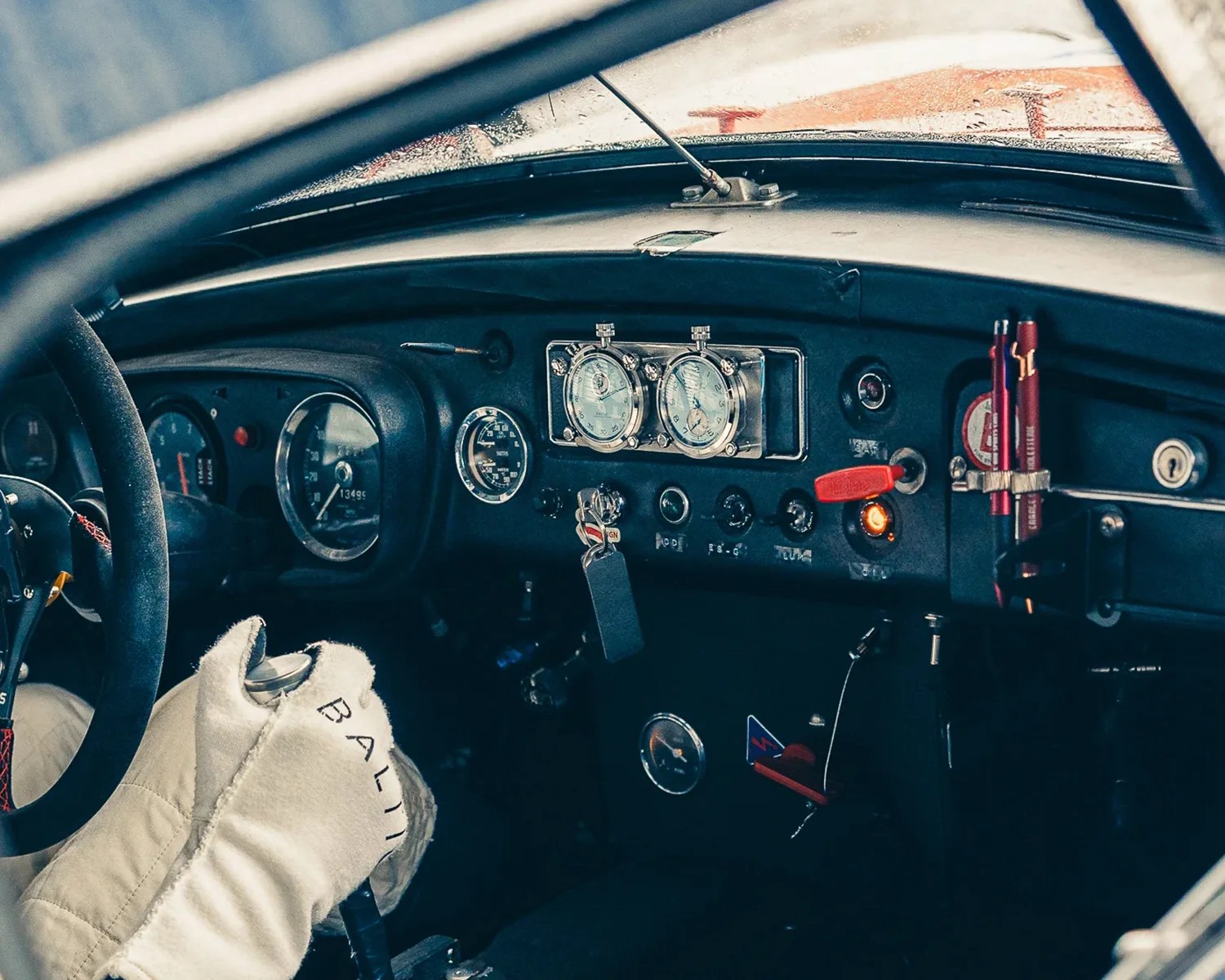 Vintage car dashboard with multiple gauges, switches, and a hand in a white racing glove on the steering wheel.