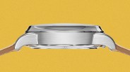 Side view of a silver stainless steel watch case with a domed crystal and tan leather straps against a yellow background.
