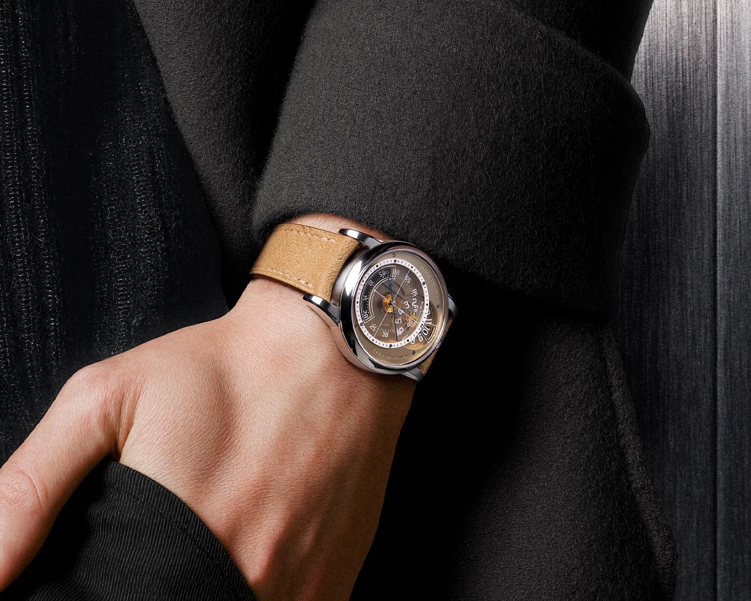 Wristwatch with a silver case, transparent dial, and tan leather strap worn on a wrist over a black coat sleeve.