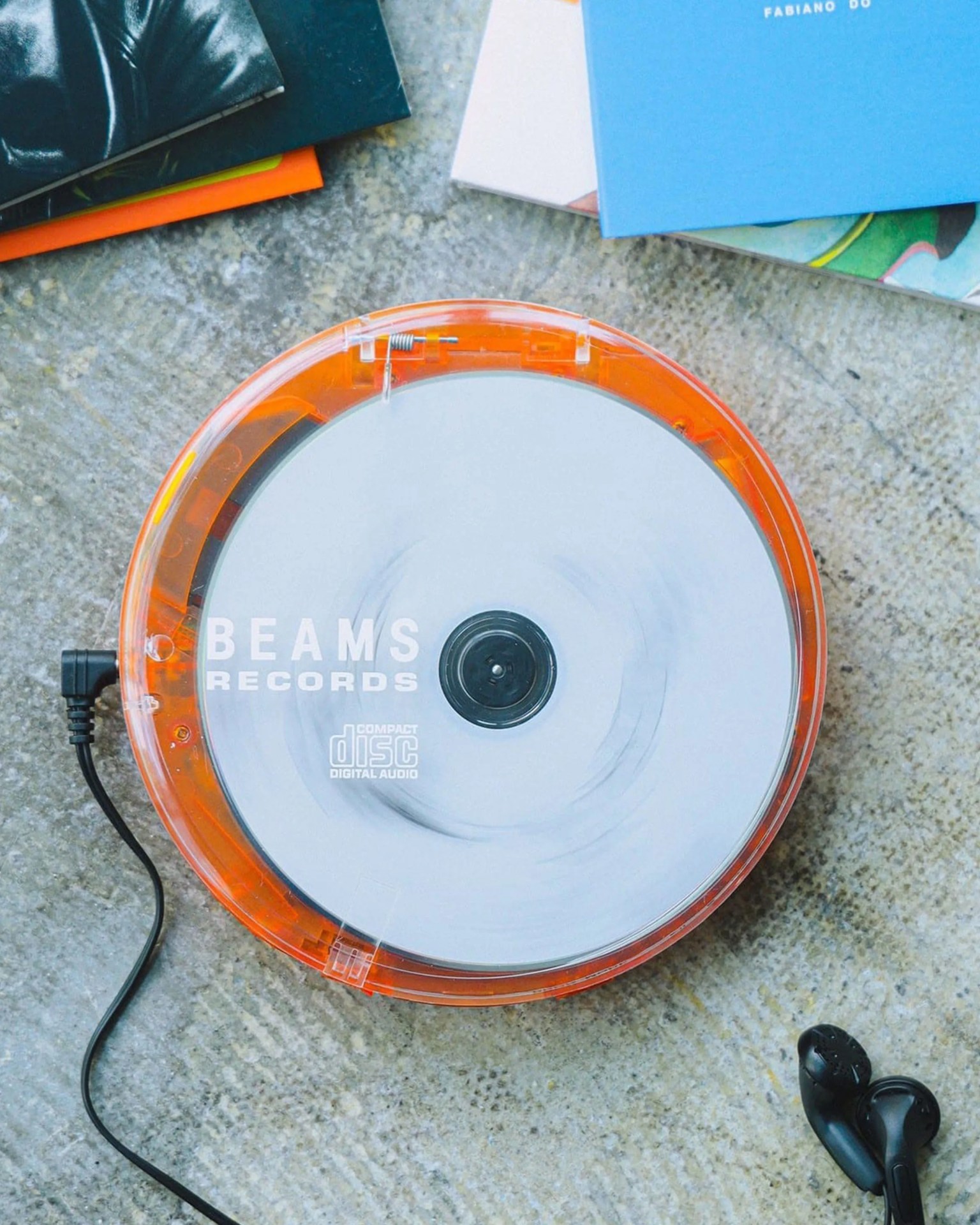 Orange transparent portable CD player with white disc labeled "BEAMS RECORDS" and black wired earbuds on a textured surface.