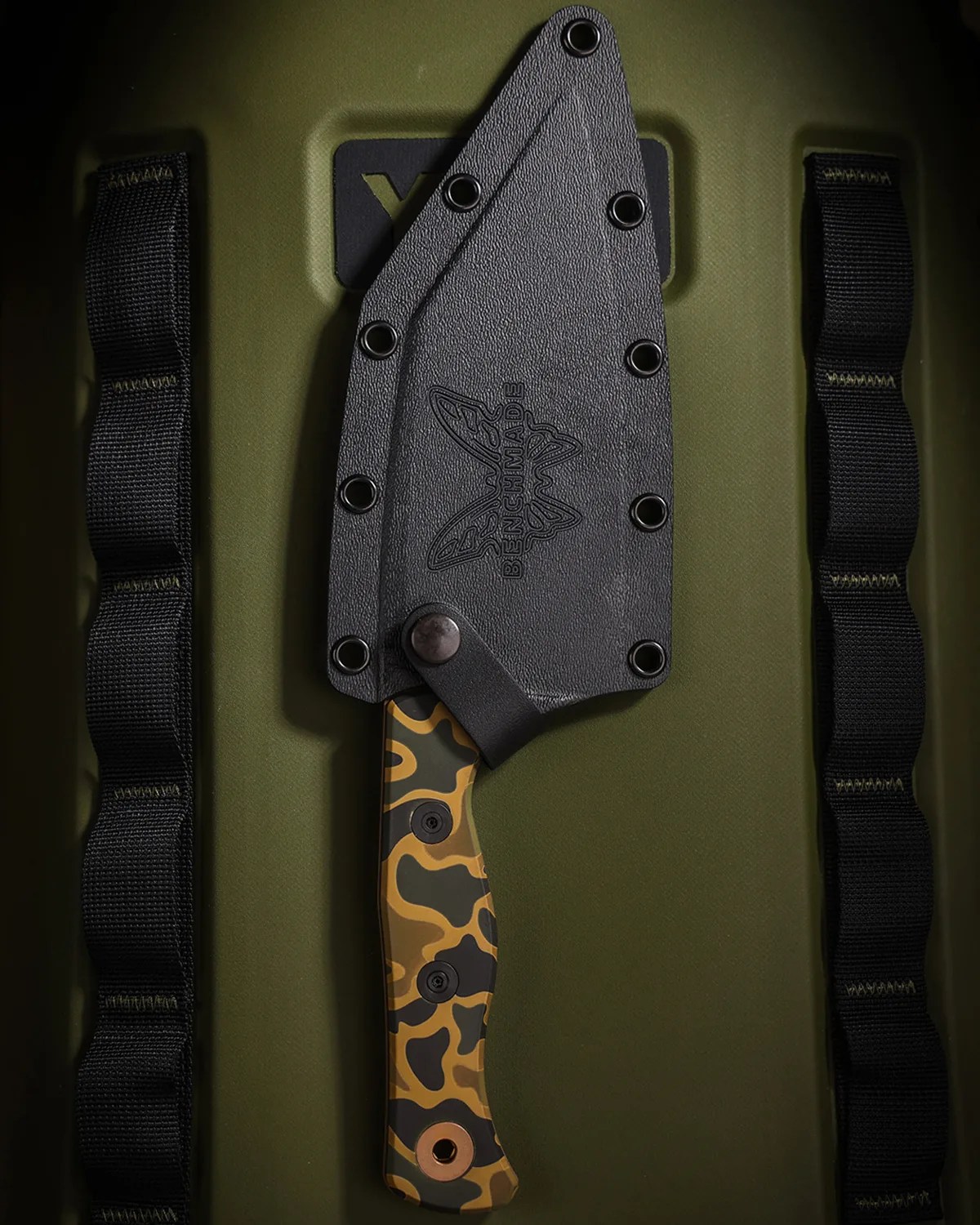 Black textured knife sheath with "Benchmade" logo covering a camo-patterned knife handle on an olive green surface.