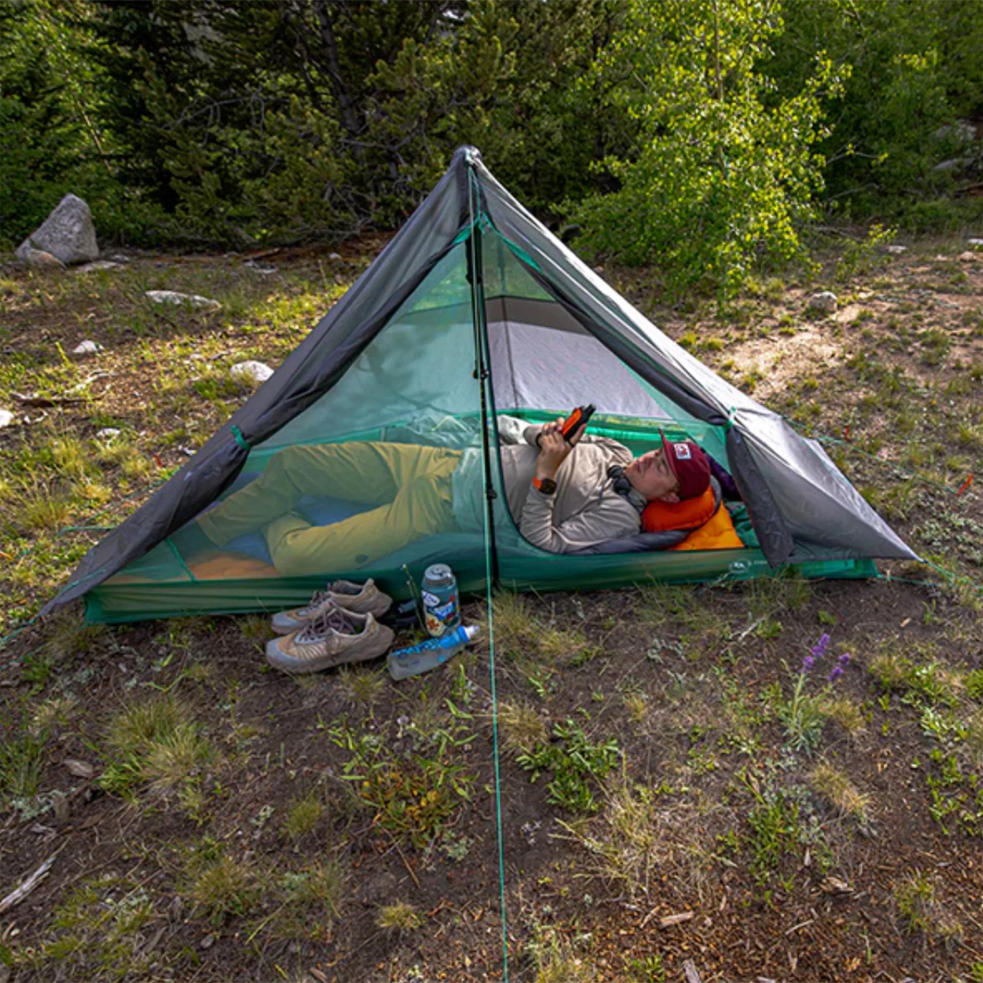 Green and gray mesh tent pitched on grass with a person lying inside using a phone.